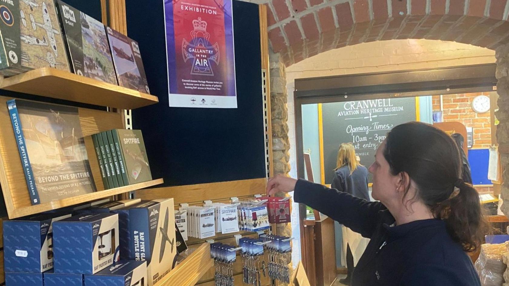 A woman facing sideways towards a wall, restocking some keychains on hooks in a shop. There are shelves with books and other items on. Another woman is writing on a blackboard in the background.