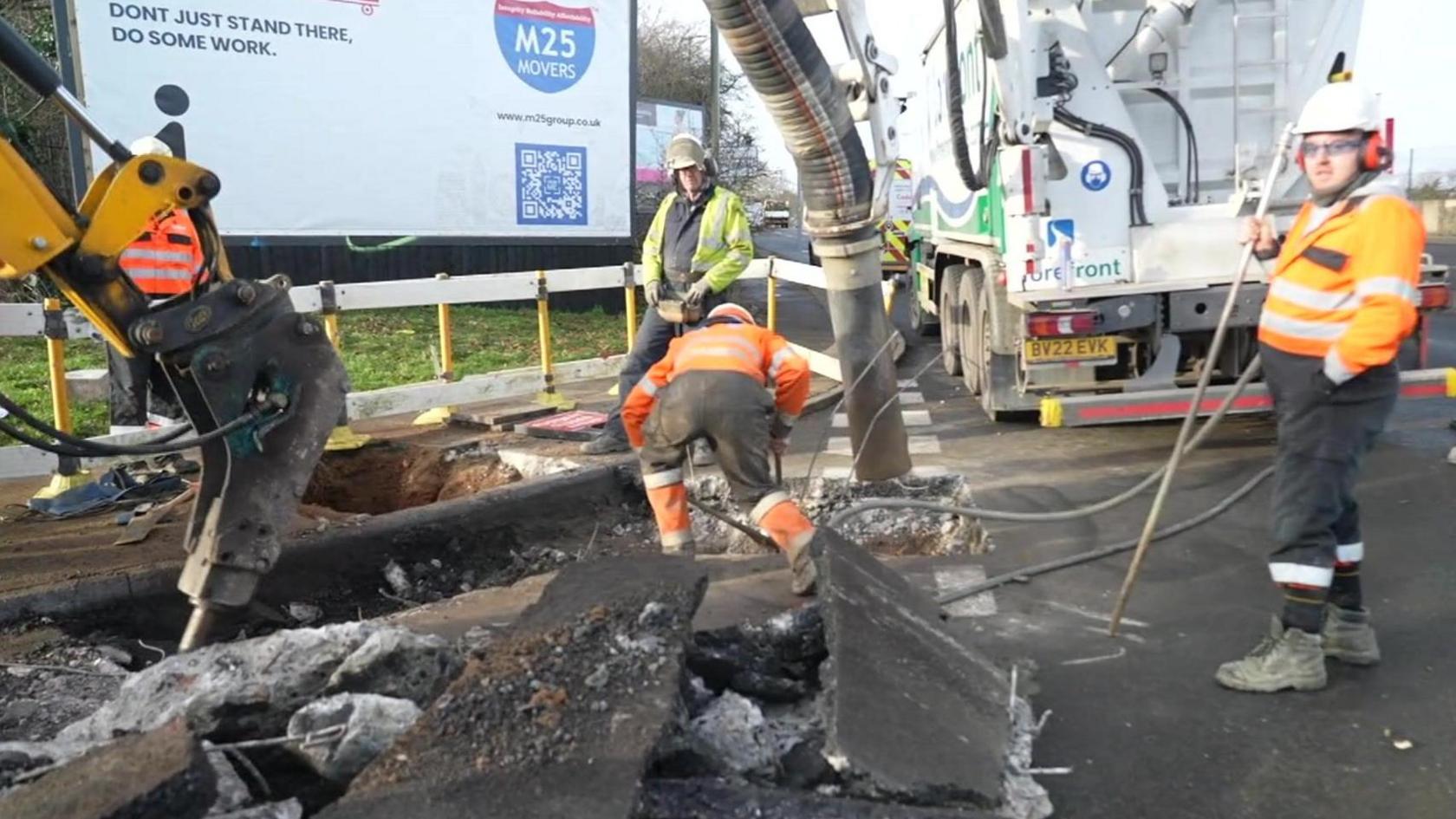 Workmen in high viz jackets and helmets working with a crane by a huge hole in the road.