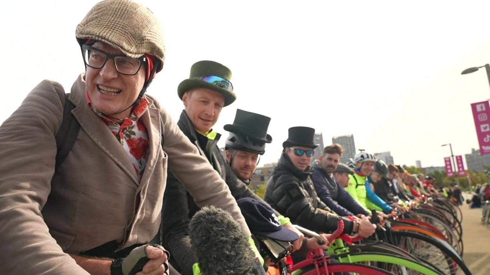 Broadcaster Jeremy Vine in a floral shirt, brown jacket, flat cap style bicycle helmet and glasses sits on a Penny Farthing in a group of riders.