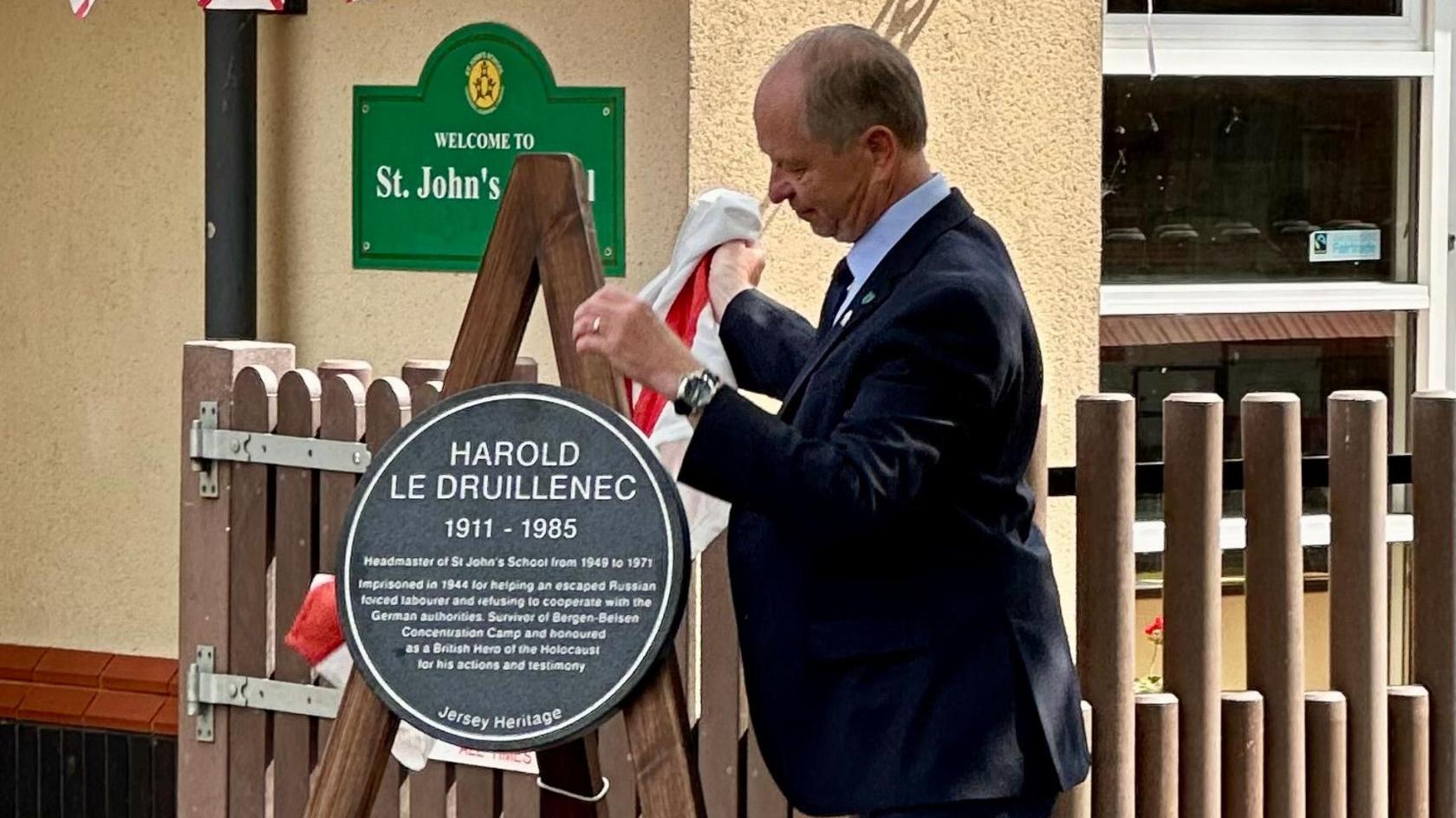 The Lieutenant-Governor removes a Jersey flag to reveal the plaque which is sitting on a wooden frame. A sign on the wall behind says: Welcome to St John's School.