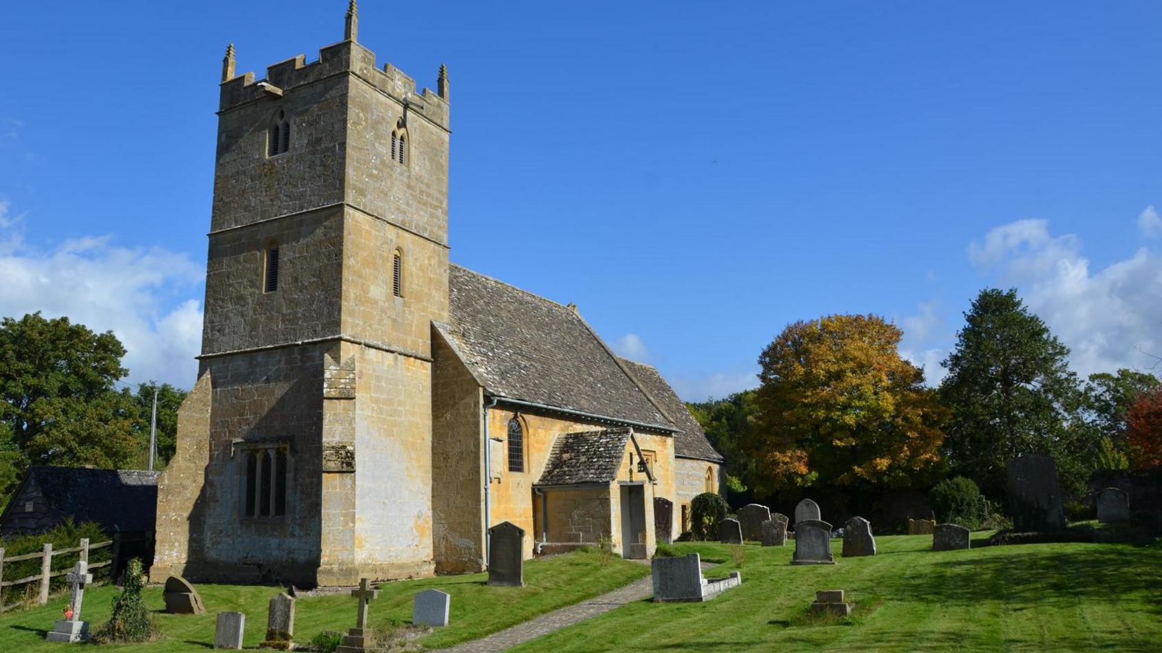 St John the Baptist Church in Wickhamford, between Broadway and Evesham, dates back to the 13th Century. The building is seen on a sunny day under a blue sky. There are headstones around it and trees in the background.