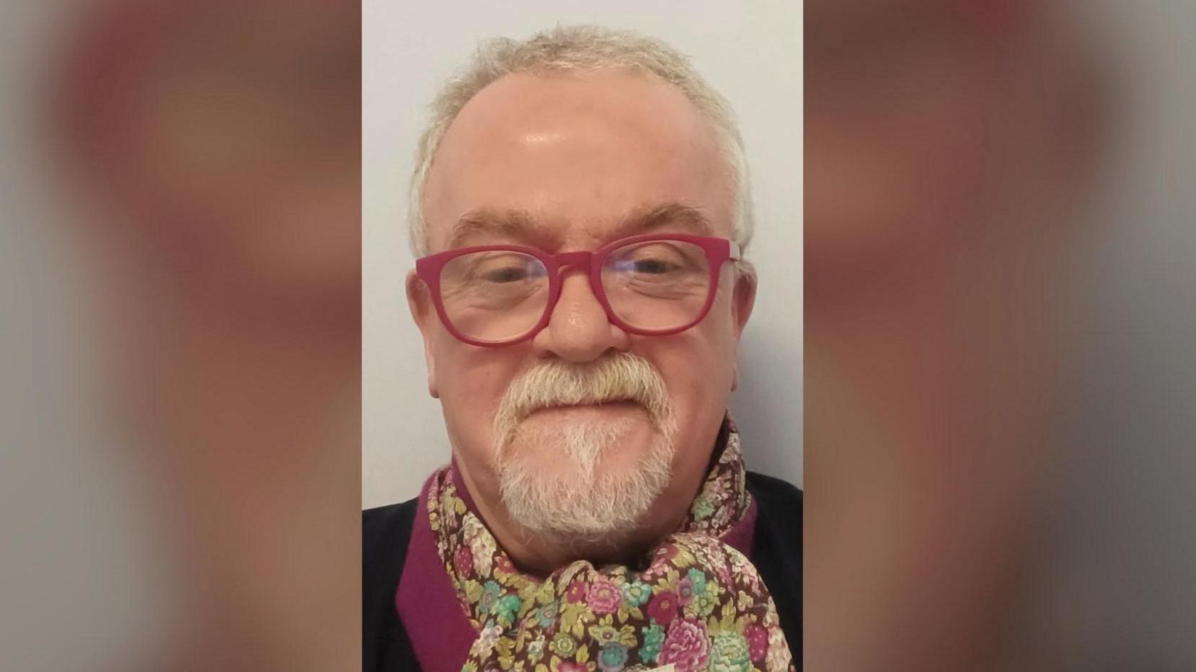 A man with white hair and a goatee looks at the camera. He is wearing pink glasses and a colourful scarf. 