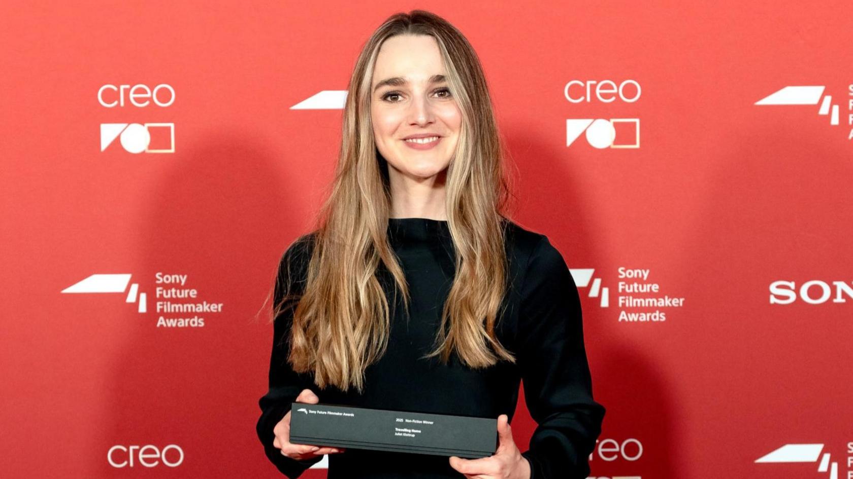 A woman in a black dress with long golden hair stands in front of a red background holding a black oblong box.