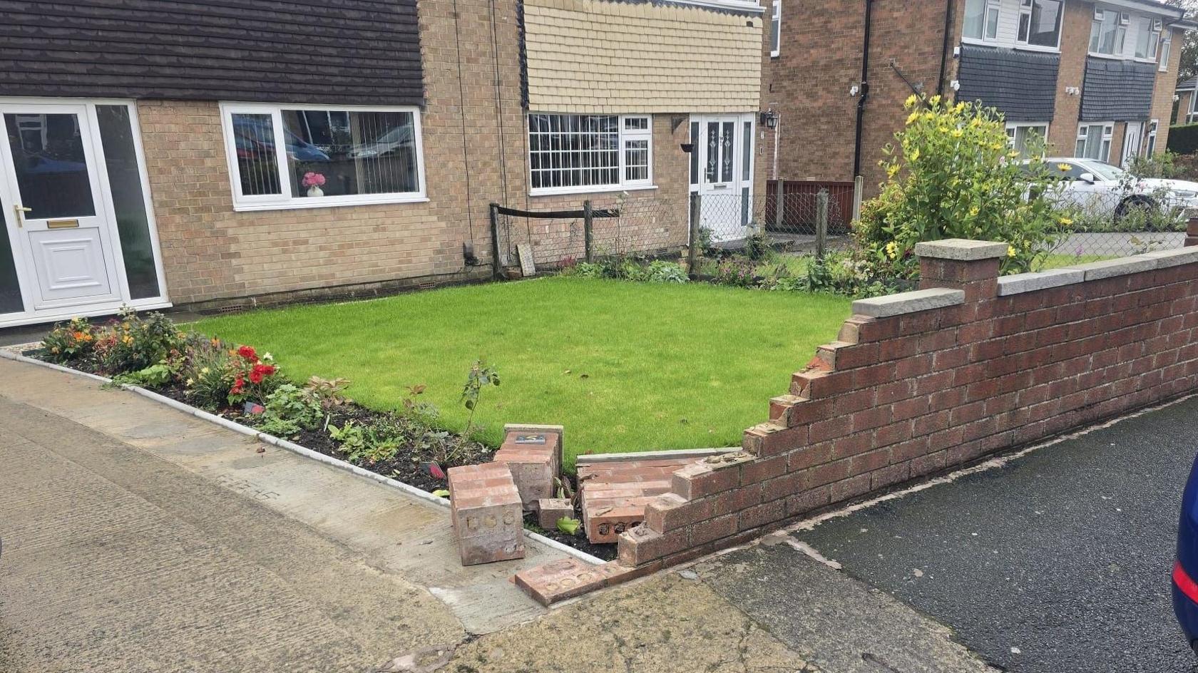 A wall outside a house. It is partly knocked down and there are bricks strewn in the driveway and front garden.