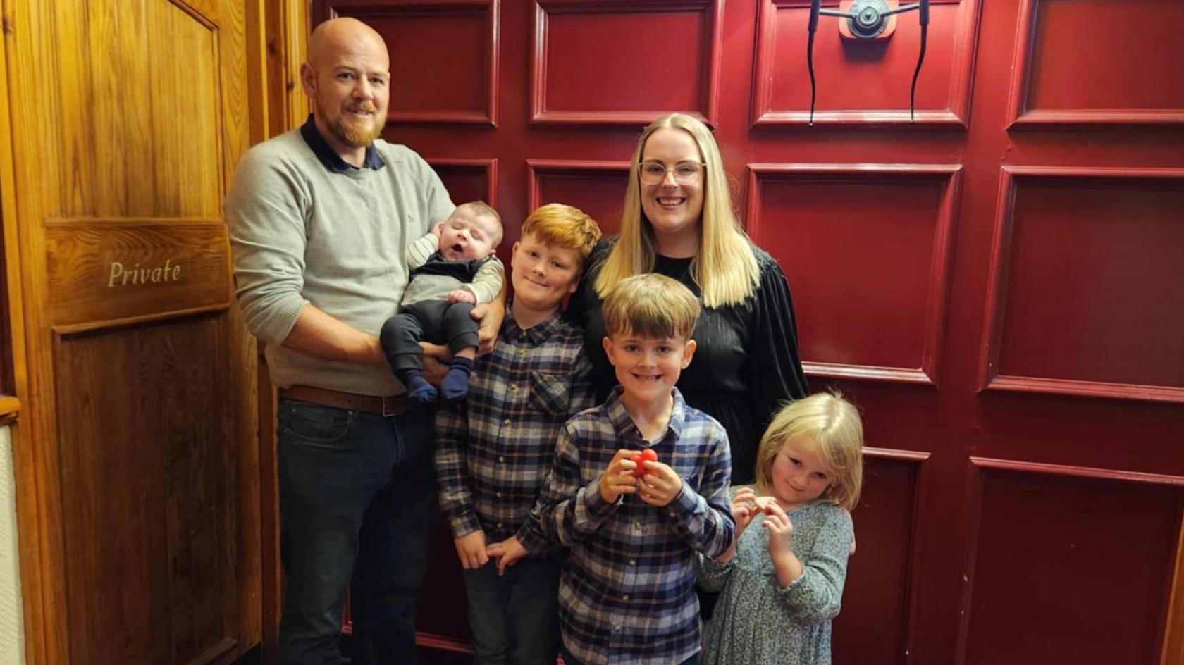 Nick and Amanda with their four children, three boys and a girl. The boys are wearing shirts and the girl wears a dress, and all are smiling in front of a red panelled wall. 