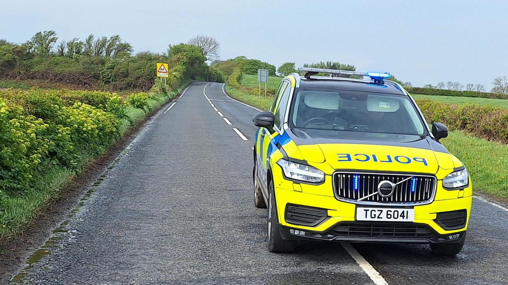 A police car is parked a country road. Green fields and hedges are on either side of the road.