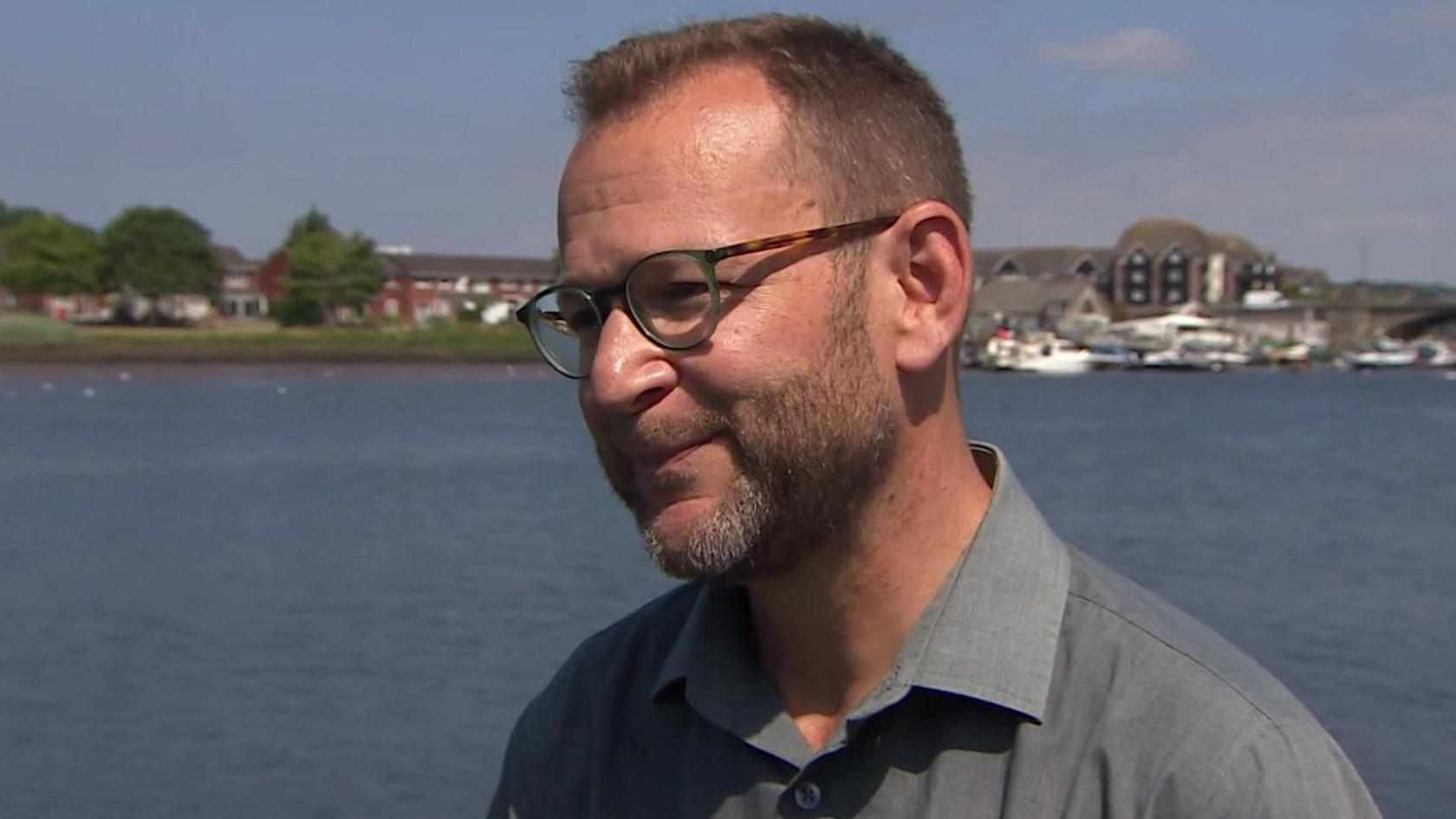 Dr Dave Rumble stands looking slightly off camera as he gives a TV interview. Behind him is the River Itchen, a large expanse of blue water with some greenery and a boathouse in the background and a few white yachts docked on the water. Dr Rumble has short light brown hair and a short beard, with tortoiseshell round thick framed glasses and a grey collared shirt.