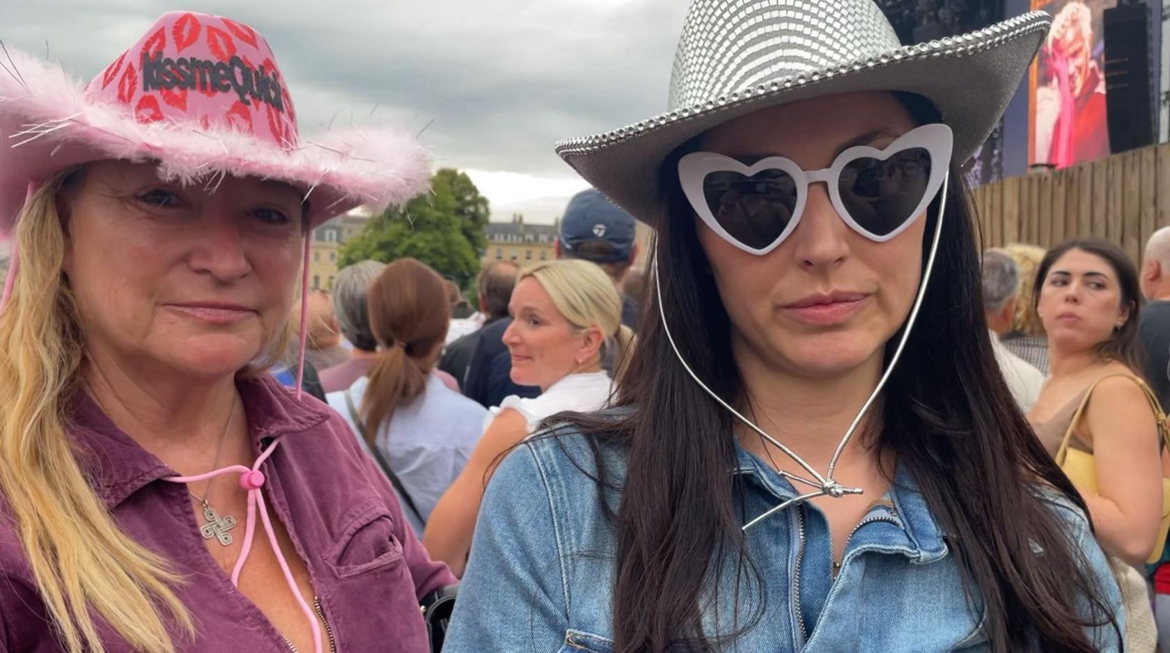 Rina standing on the right wearing a denim shirt, a disco ball cowboy hat, and white framed sunglasses shaped like a heart. Beside her is her aunt Stella, wearing a pink jumpsuit and pink cowboy hat decorated with lips and a fluffy rim. Behind them you can see the main stage and a large crowd of people.