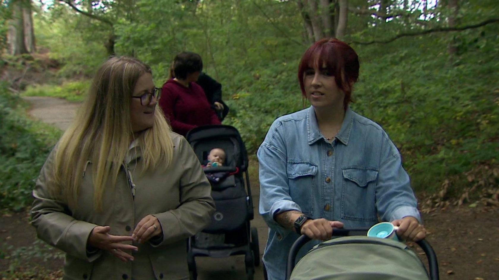 A woman with long blonde hair and a beige coat walking next to a woman with red head and a push chair wearing a denim jacket in woodland.
A woman with brown hair is behind the pair wearing red with a pushchair with a small baby