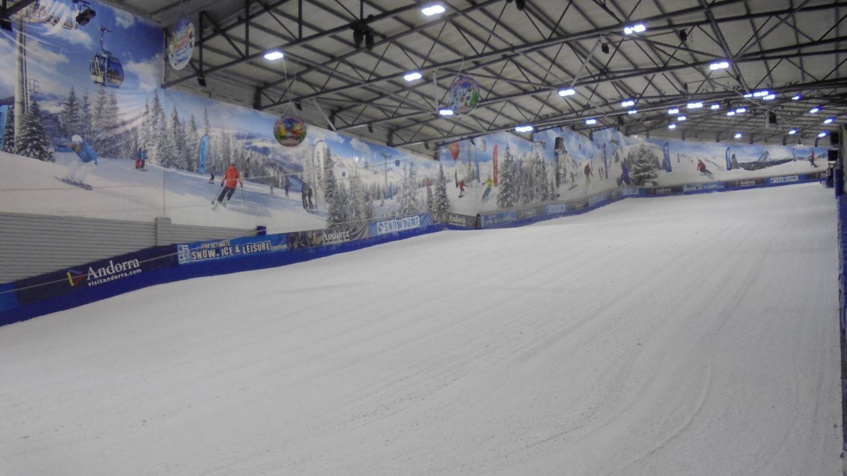 An indoor ski slope with lights on the roof and advertising boards along the walls that show people skiing. 