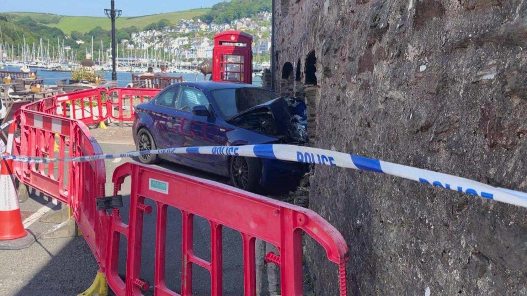 A blue car with the bonnet crushed against a building with police tape around the scene and orange cones cordoning it off.