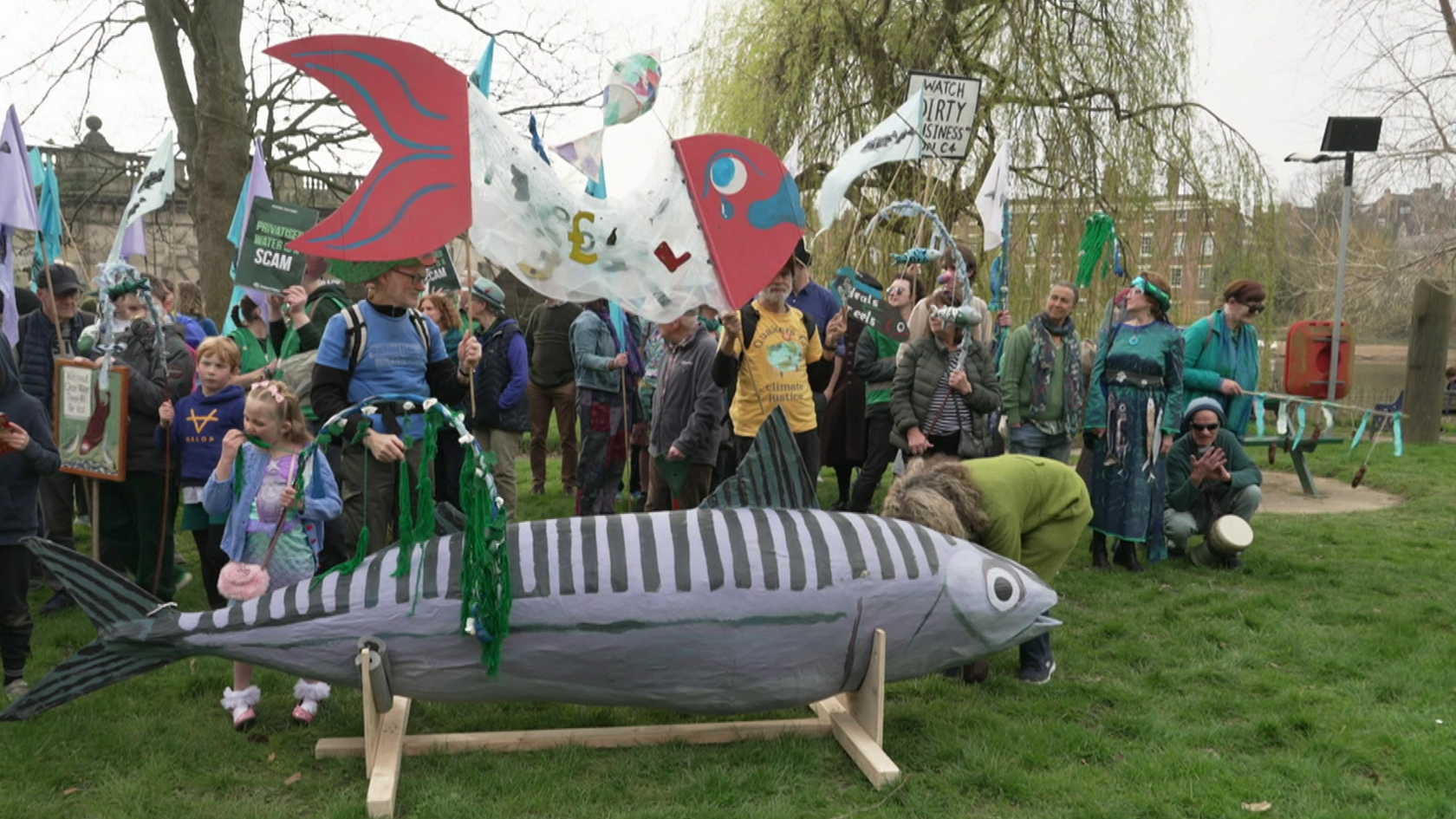 A large grey model fish can be seen in front of about 40 to 60 people, some waving banners and placards, by the river. They are standing on grass.