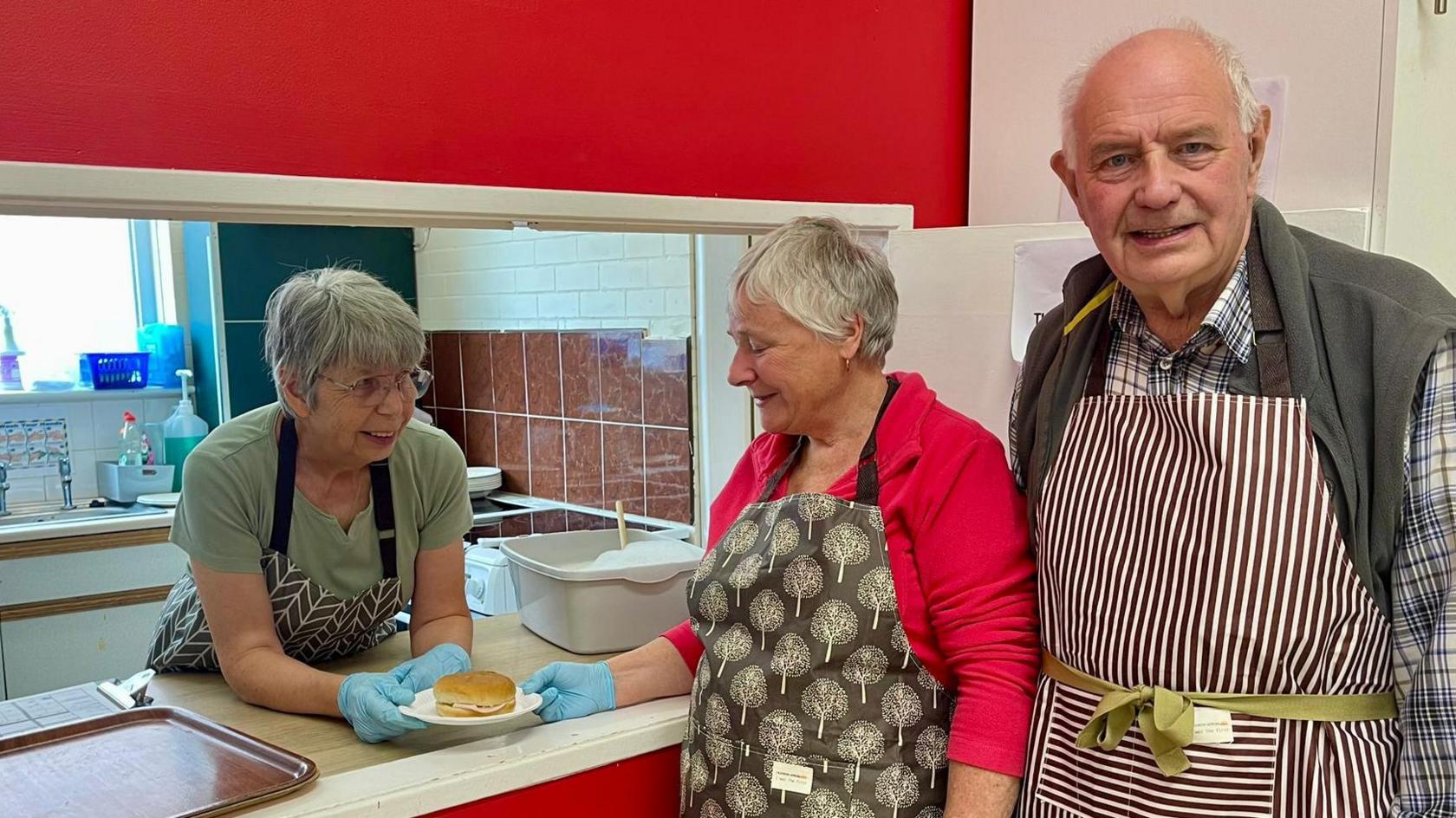 Two women and a man dressed in aprons prepare sandwiches inside a church kitchen.