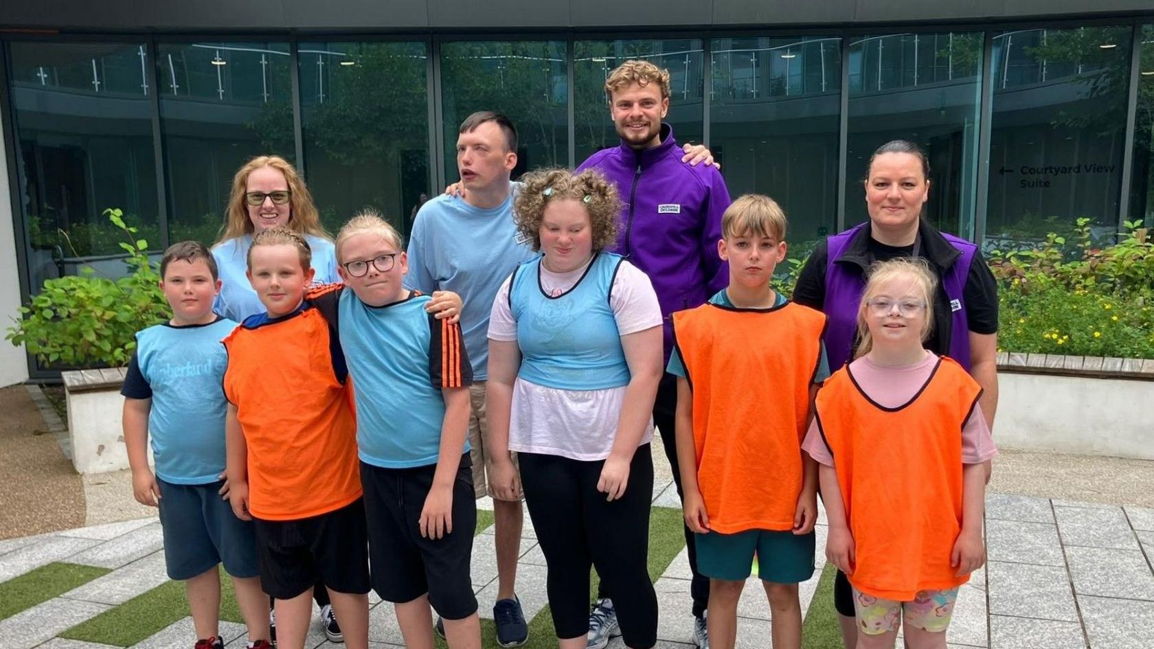 A group of people wearing sports bibs are standing together for a posed photo. There are three people wearing orange bibs, as well as four people wearnig blue bibs and two people wearing purple jackets.