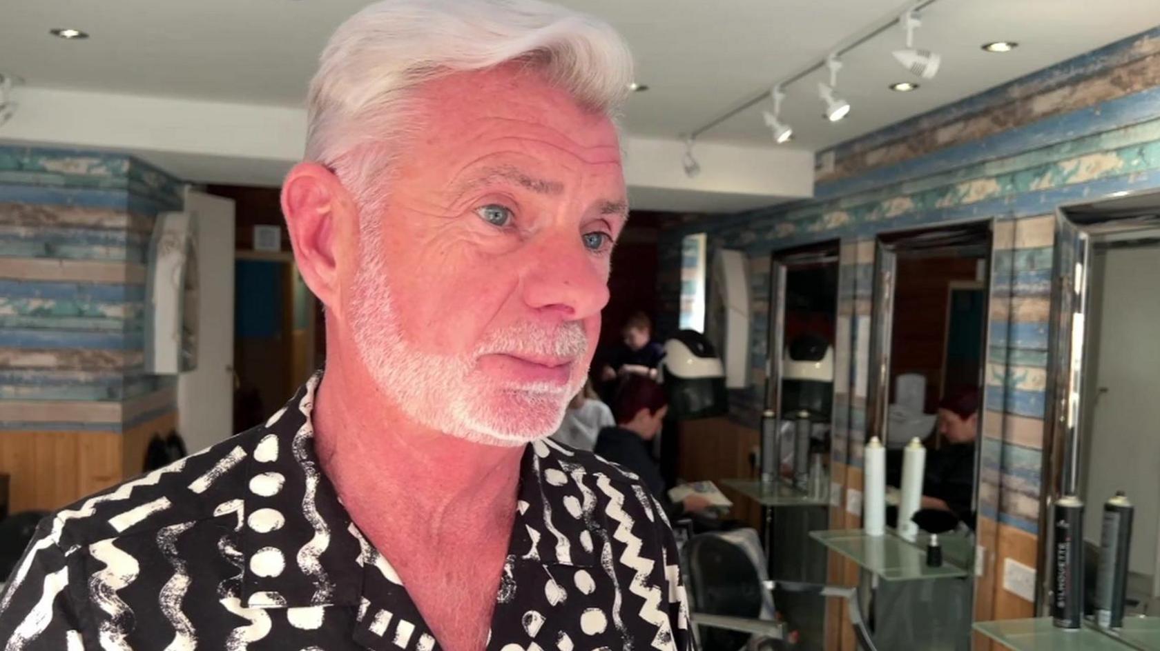 Michael Elliott-Jones standing in his salon. He has short white hair and a neatly-trimmed full beard and is wearing a black shirt with white zig-zag and dot patterns