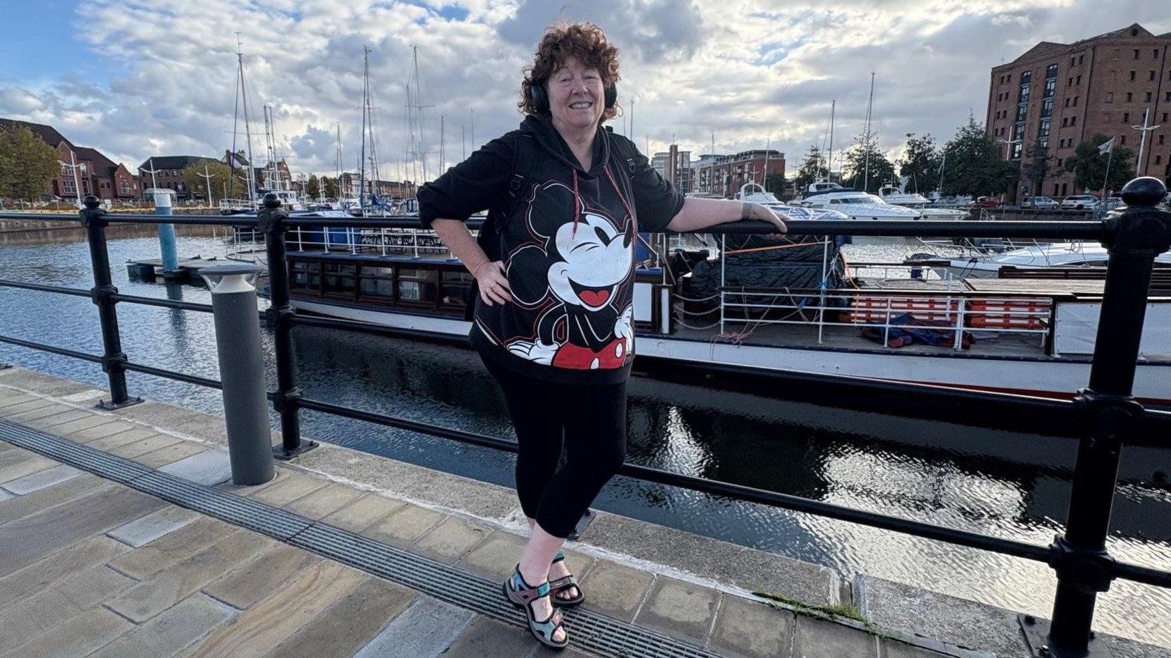 A woman in her 60s with brown curly hair is standing on path with a marina and yachts in the background. She is wearing a black T-shirt with a picture of Micky Mouse on it