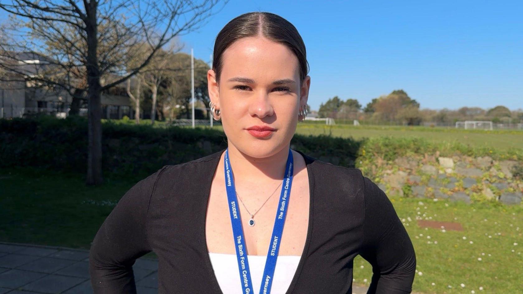 A teenage girl with a slick back brunette pony tail. She is wearing a white top with a black cardigan. She has a blue student lanyard on and big hoop earrings on 