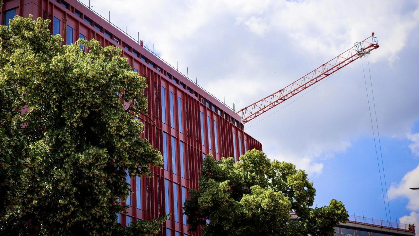 The side of one of the buildings, taken from street level. Large trees are in front of part of the development, with a crane overhead. 