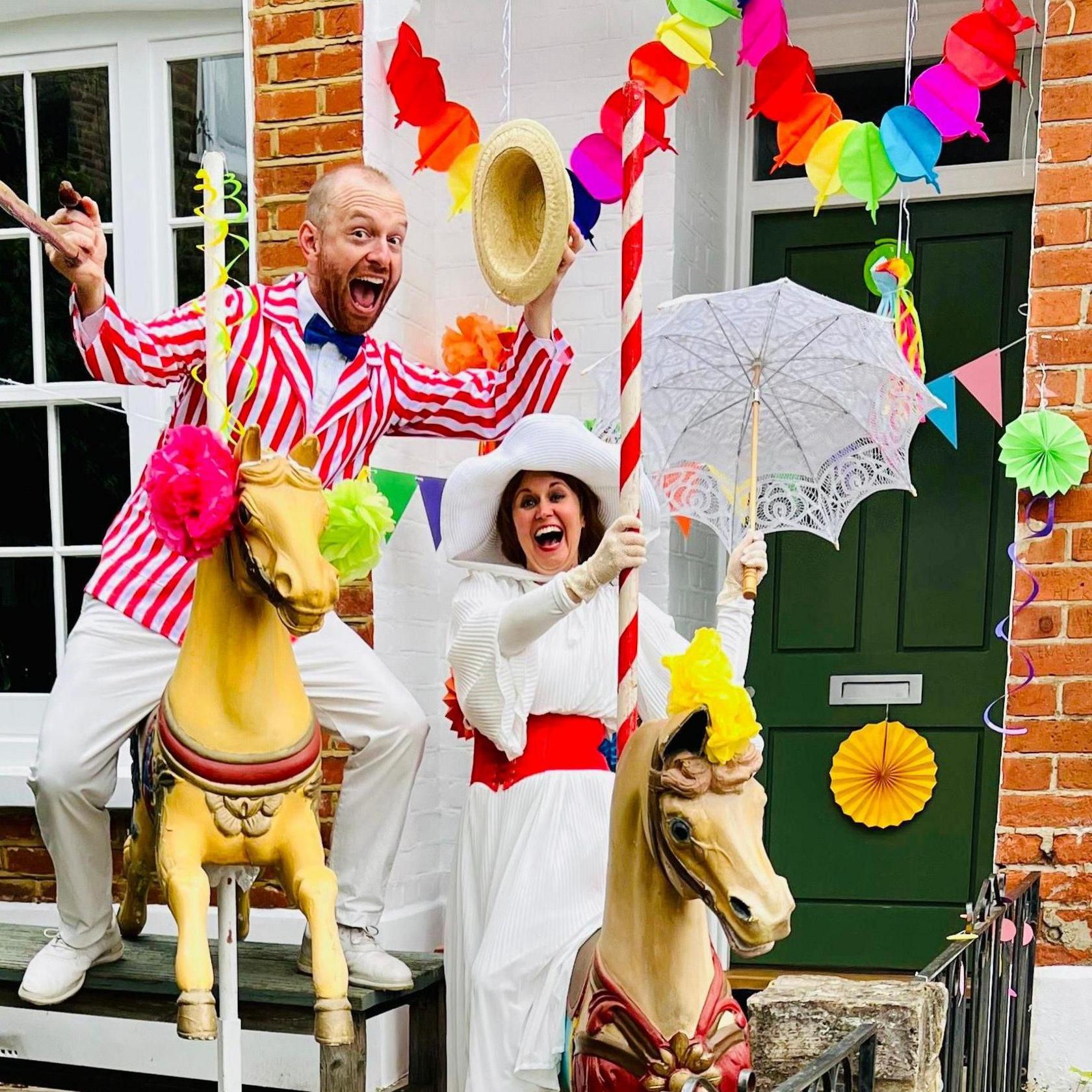 Tom, dressed as Bert in a red-and-white striped blazer and straw hat, poses on a carousel horse. His wife Beth, dressed as Mary Poppins in a white dress and wide-brimmed hat, holds a lace parasol. Their house is decorated with colourful paper garlands.