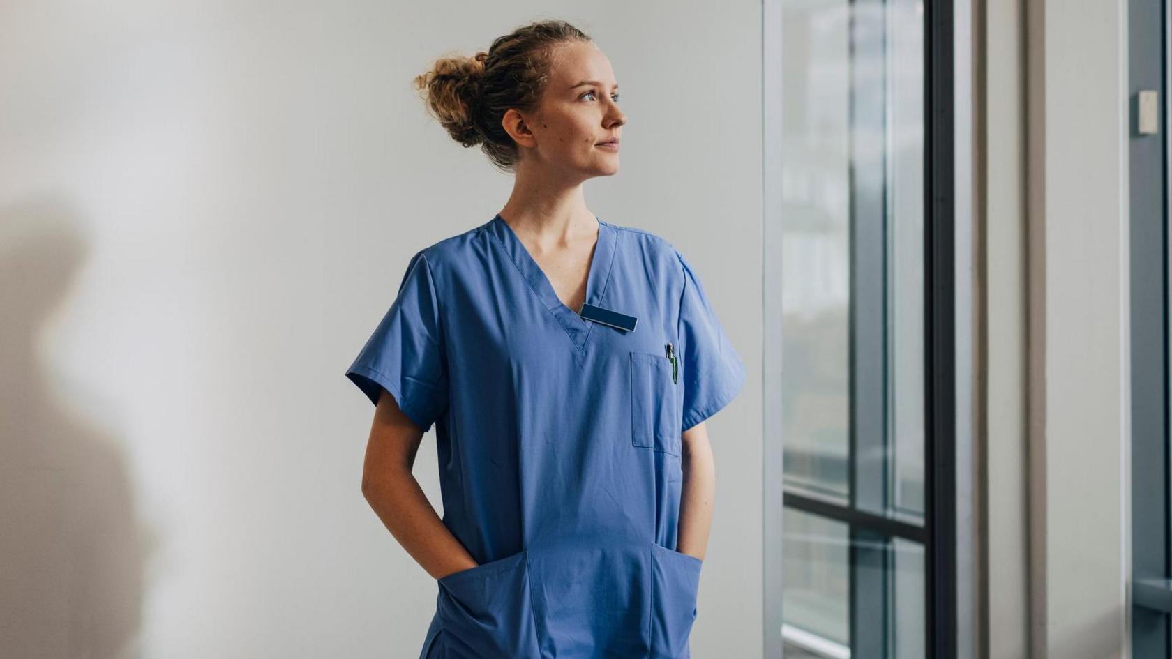 A female media stands in blue scrubs and looks out the window