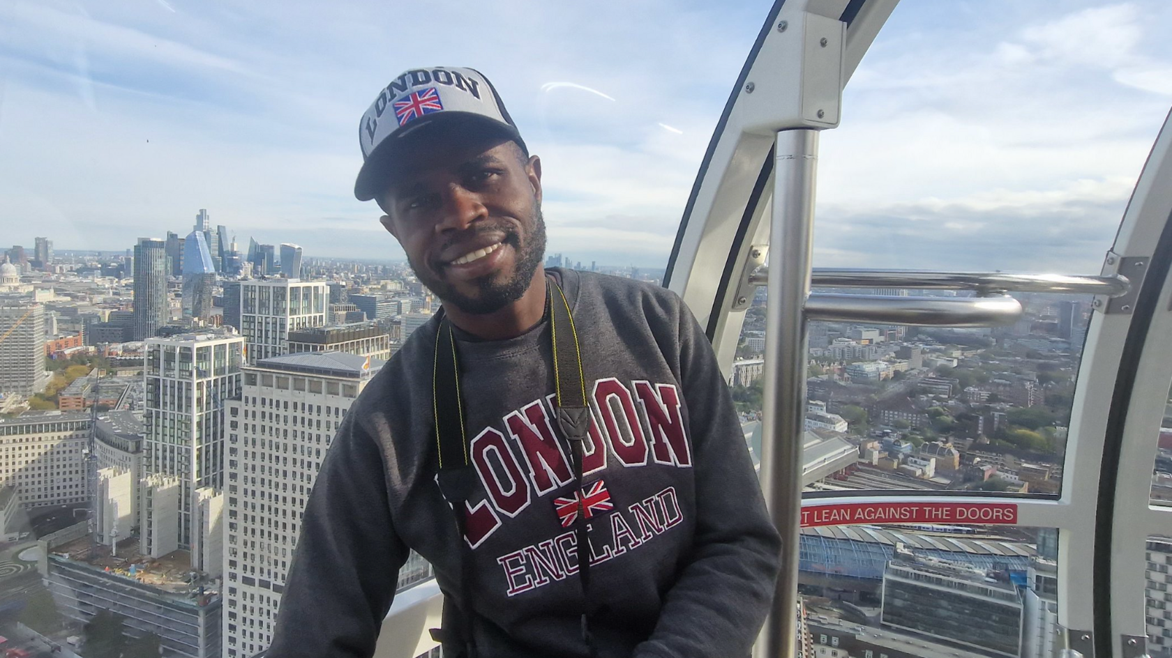 Yostin Mosquera is standing in a pod on the London Eye tourist attraction, smiling at the camera. He is wearing a jumper and a hat, both with "London" written on them. In the background is the London skyline with several skyscrapers in the distance.