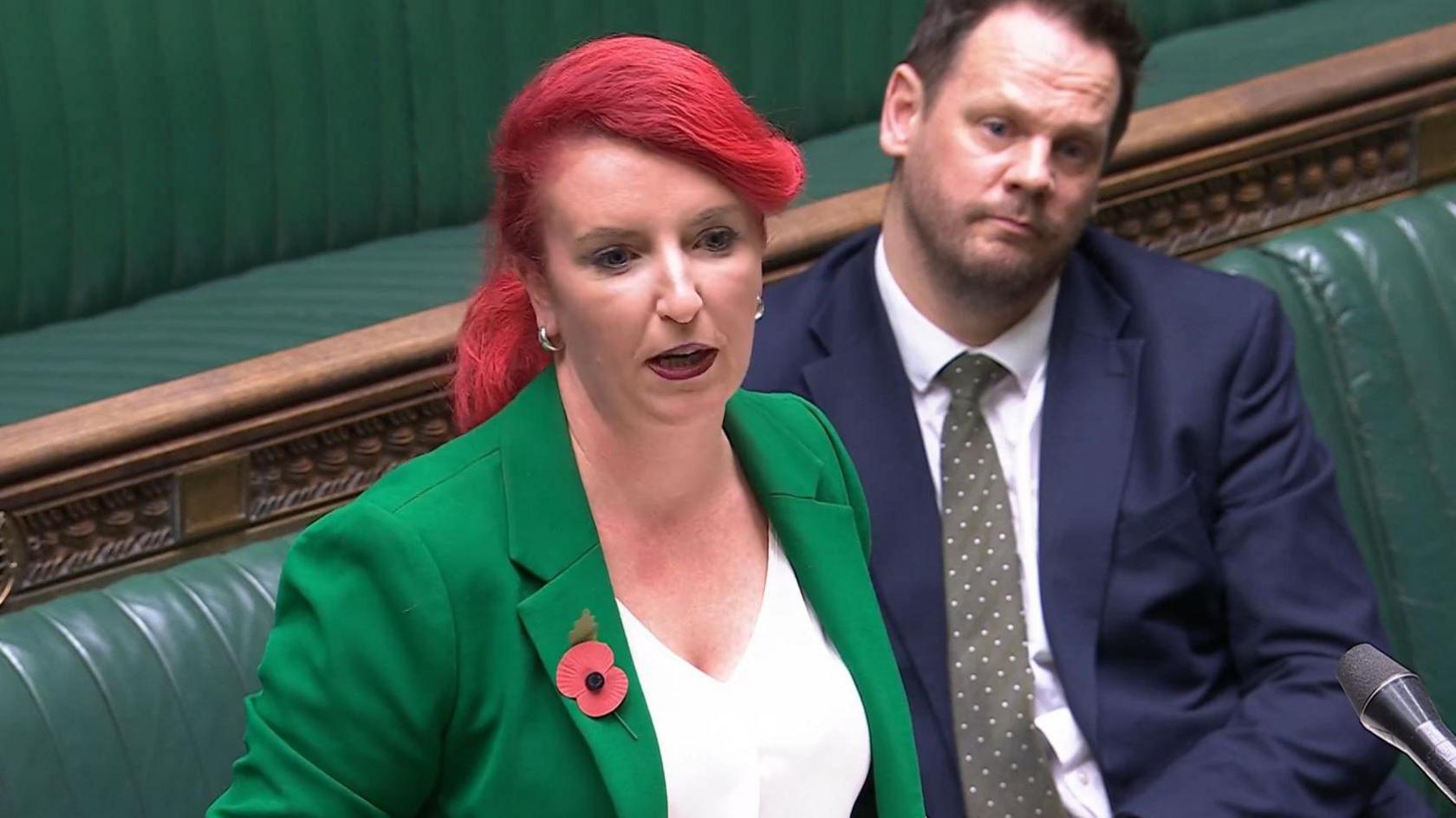 Transport Secretary Louise Haigh speaking at the Houses of Parliament. She is standing up. She is wearing a white top and a green blazer. A man in a dark blue suit is sitting behind her.