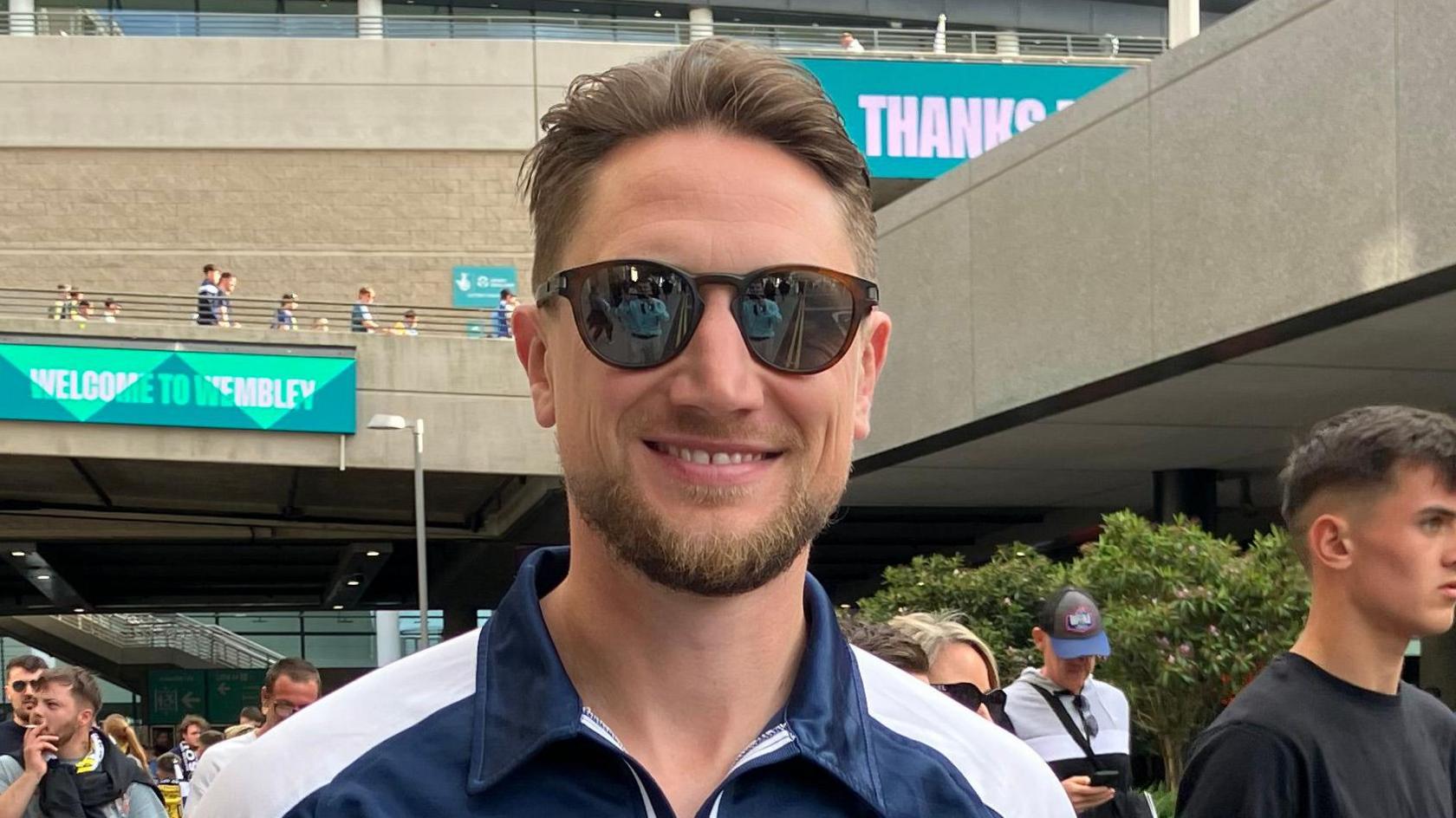 David Bonnett wearing sunglasses and a navy Southend top. The stadium and lots of fans are behind him.