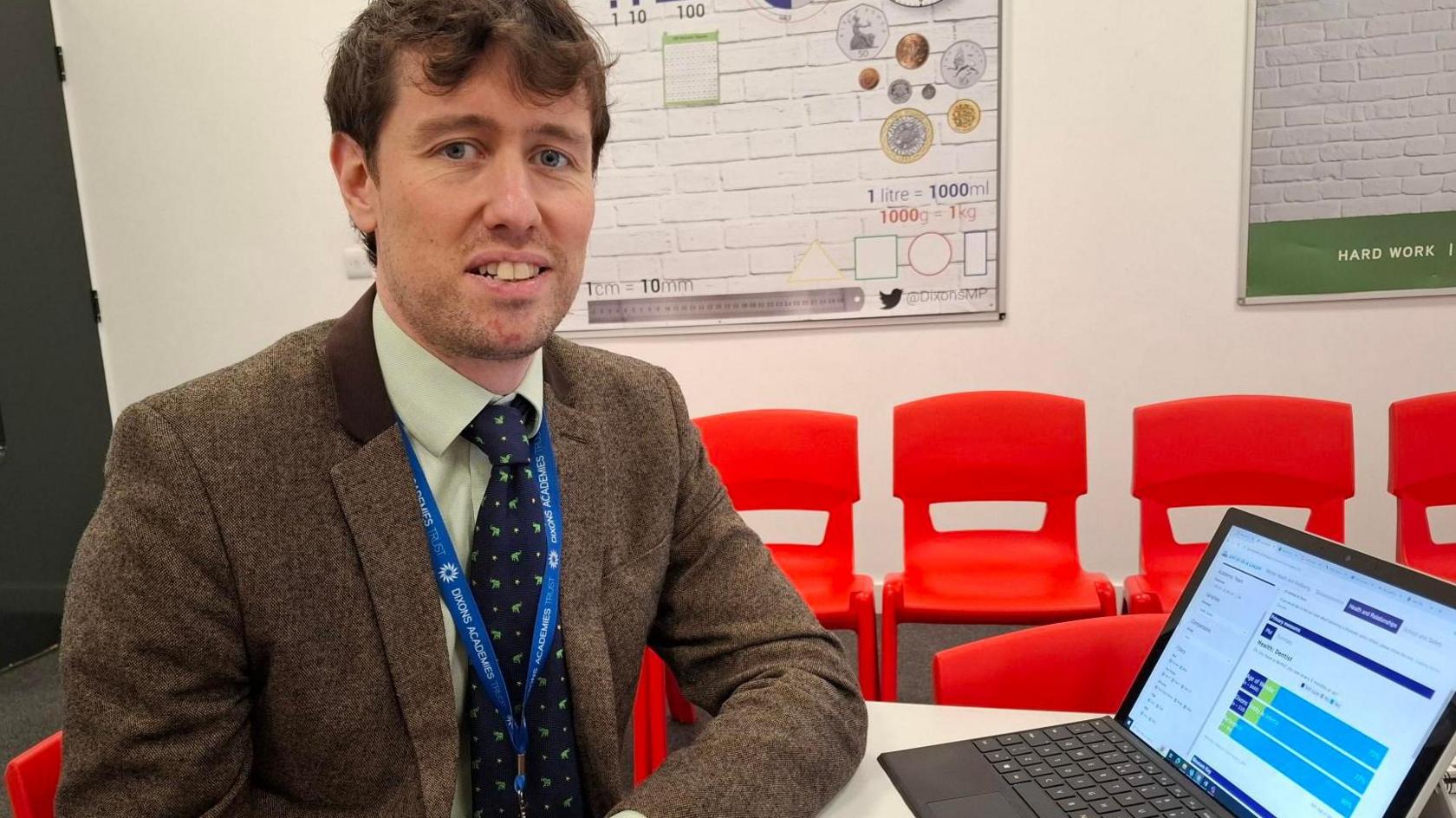 A man with brown hair wearing a brown blazer, green and white shirt and navy and green tie sits at a table. He has an open laptop in front of him with a graph on the screen. A row of bright red chairs lines the wall behind him. On the wall there is an educational poster with numbers and coins on it.