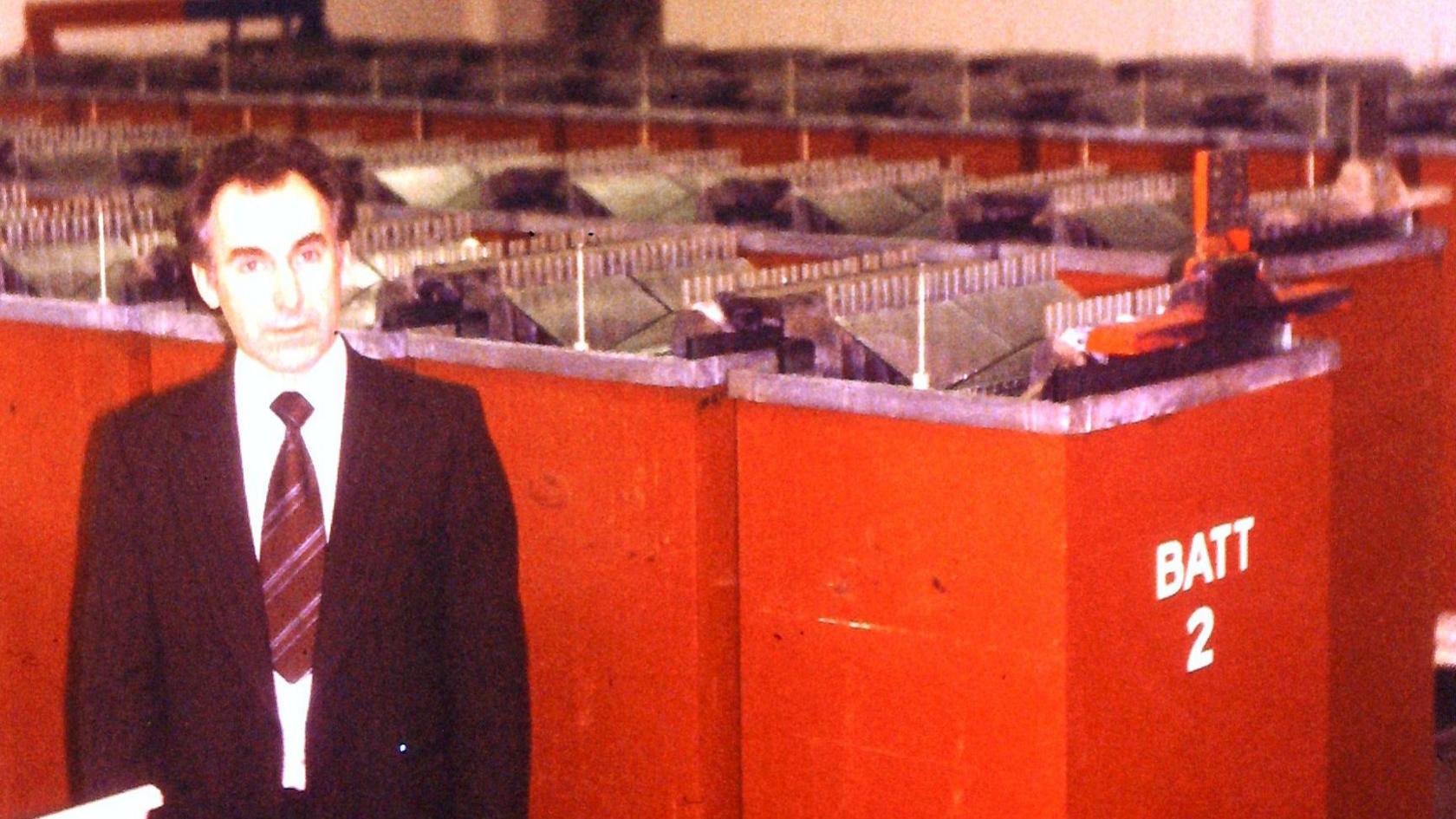 An old colour photo of a man in a black suit with a red tie. He is stood in front of rows of red boxes filled with electrical cables.