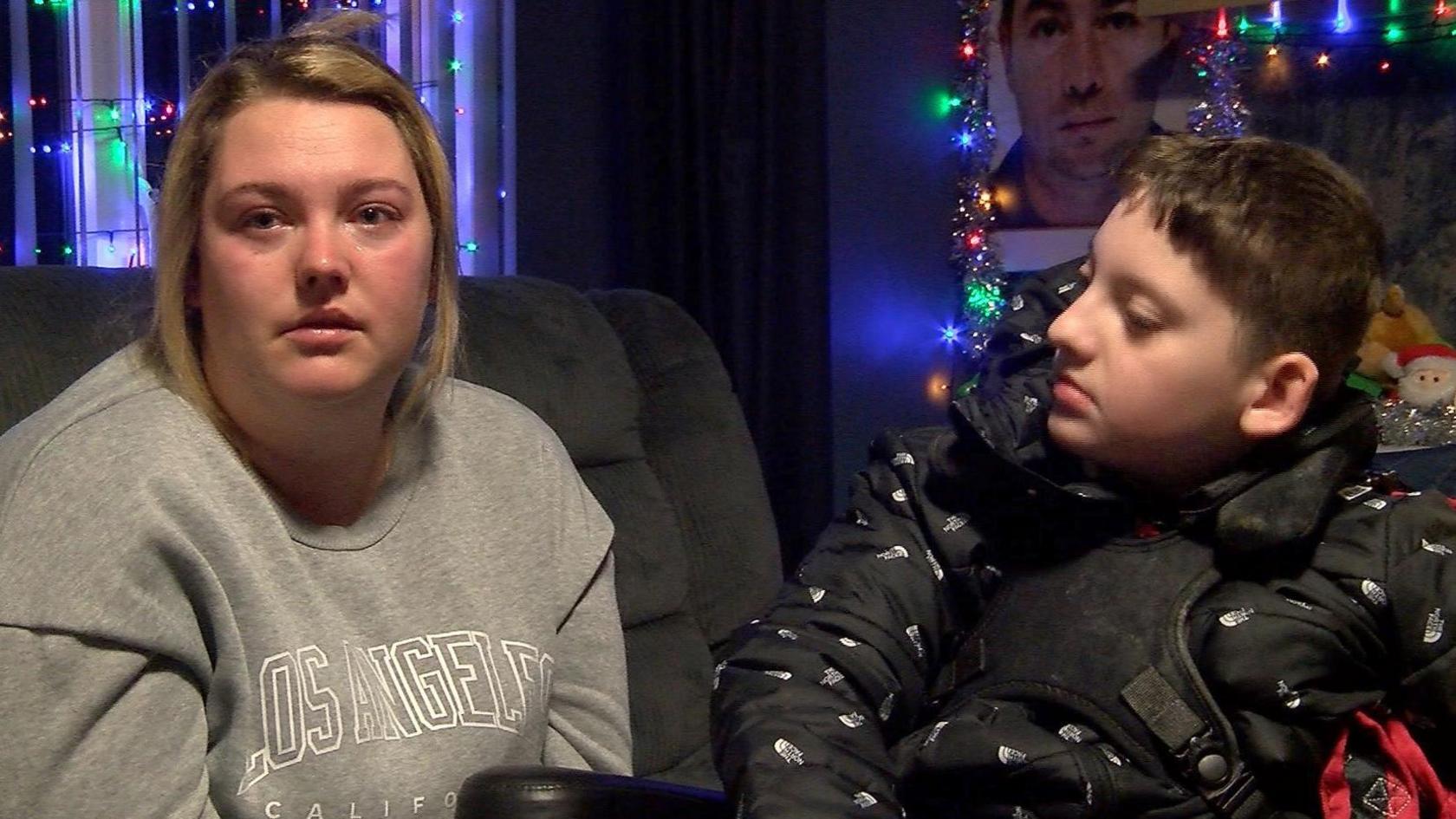 A young woman with cropped brown hair and wearing a grey sweatshirt is seen sitting on a grey sofa. To her left, her son sits in a wheelchair. He is a  12-year-old boy who has brown hair and is wearing a black coat. He is looking towards his mother. In the background, multicoloured fairy lights decorate the walls.