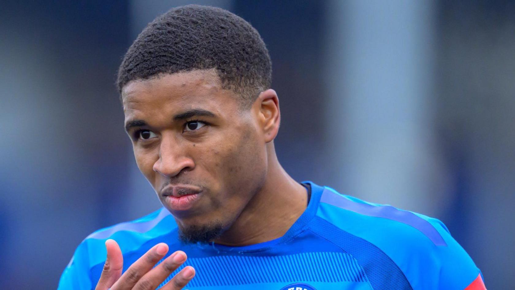 Montel McKenzie wearing the blue Billericay Town top. He looks focused and is holding his hand towards his face. He has short black hair.