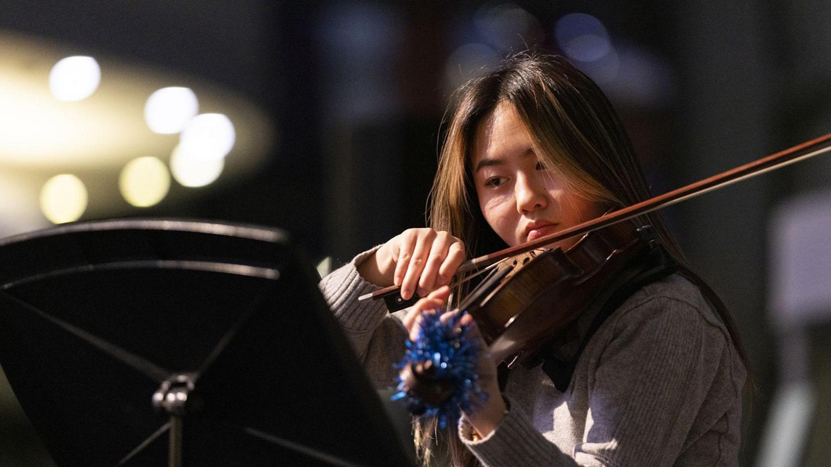 A young violinist is playing on stage. She has brown hair and is wearing a grey jumper. She is reading sheet music from a stand. Blue tinsel is tied to the head of her violin.