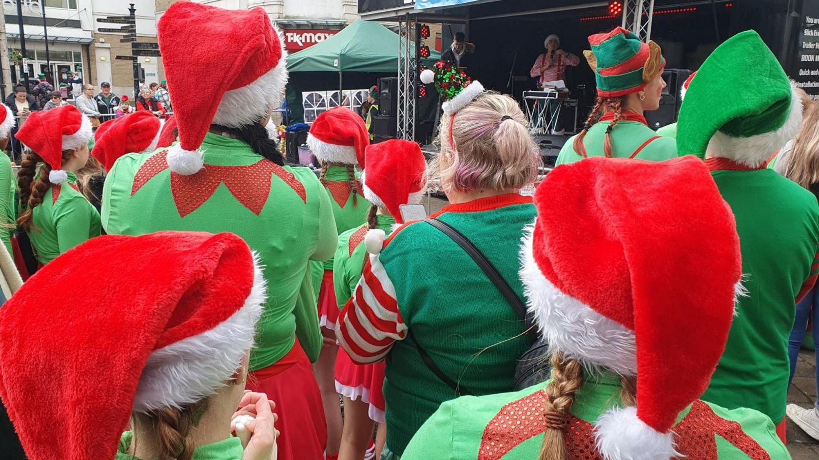 The rear view of a crowd, all dressed as elves, watching performers on an outdoor stage in a shopping precinct.