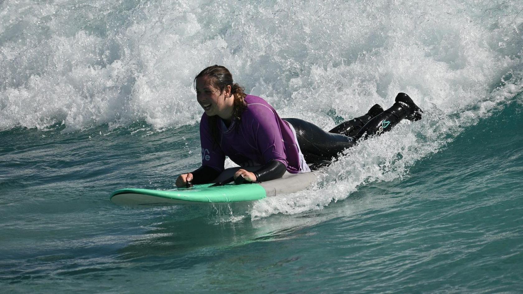 Kay Millar surfing on a wave. She is wearing a purple rash vest on top of a black wetsuit. She is surfing on a green and grey surfboard.