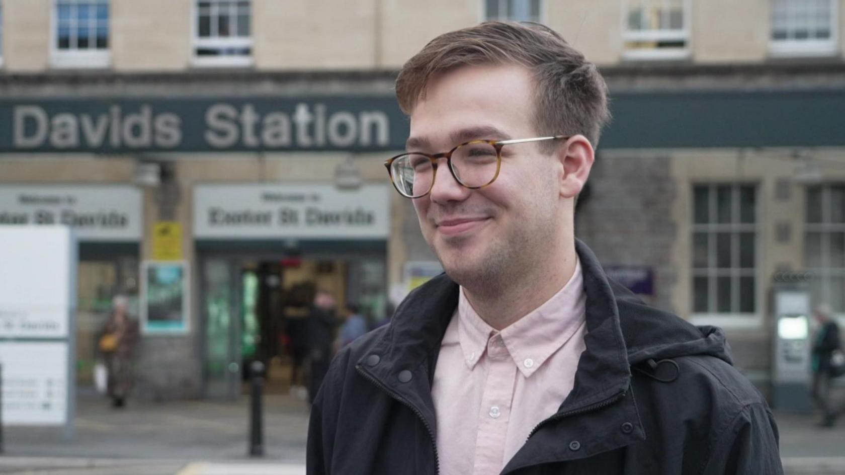A man wearing a pink shirt and a black coat looking towards the camera in front of a train station. He is wearing glasses and is smiling.