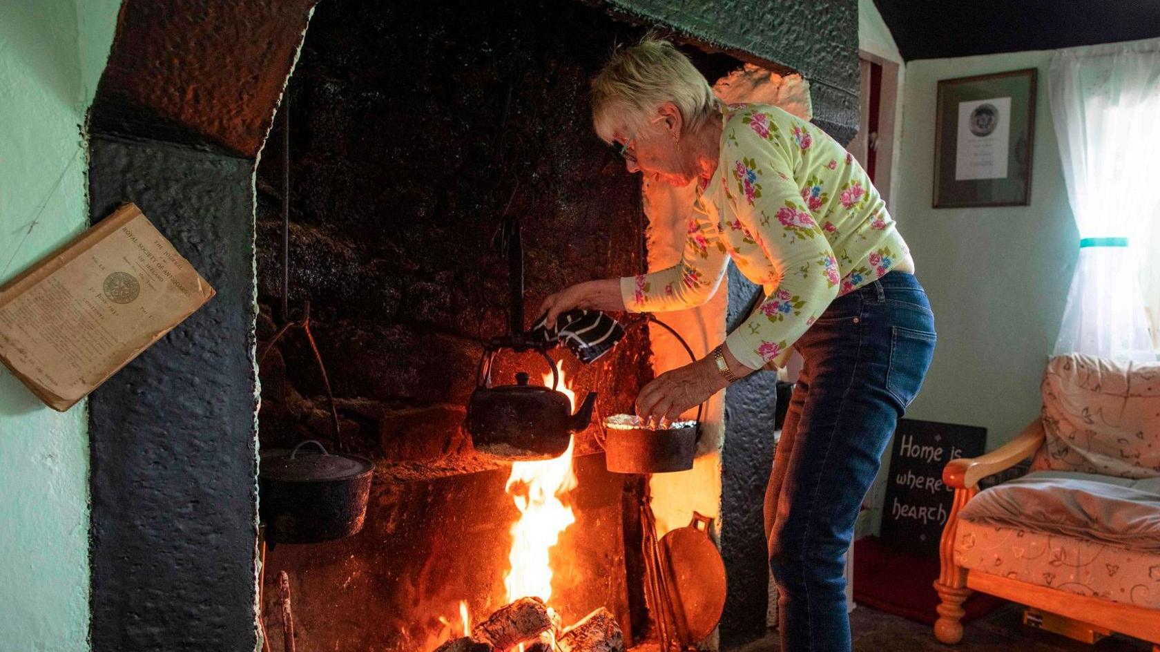 Margaret Gallagher heating a kettle of water at her large open fireplace in Mullylusty Cottage near Belcoo in 2019.  She has short, grey hair and glasses and is wearing a yellow floral T-shirt and blue jeans.  