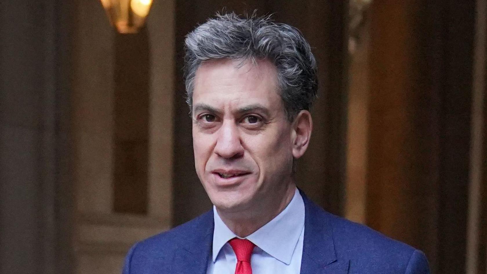 Ed Miliband is pictured smiling at the camera. He has short black and grey hair. He is wearing a dark blue suit with a light blue shirt and red tie.