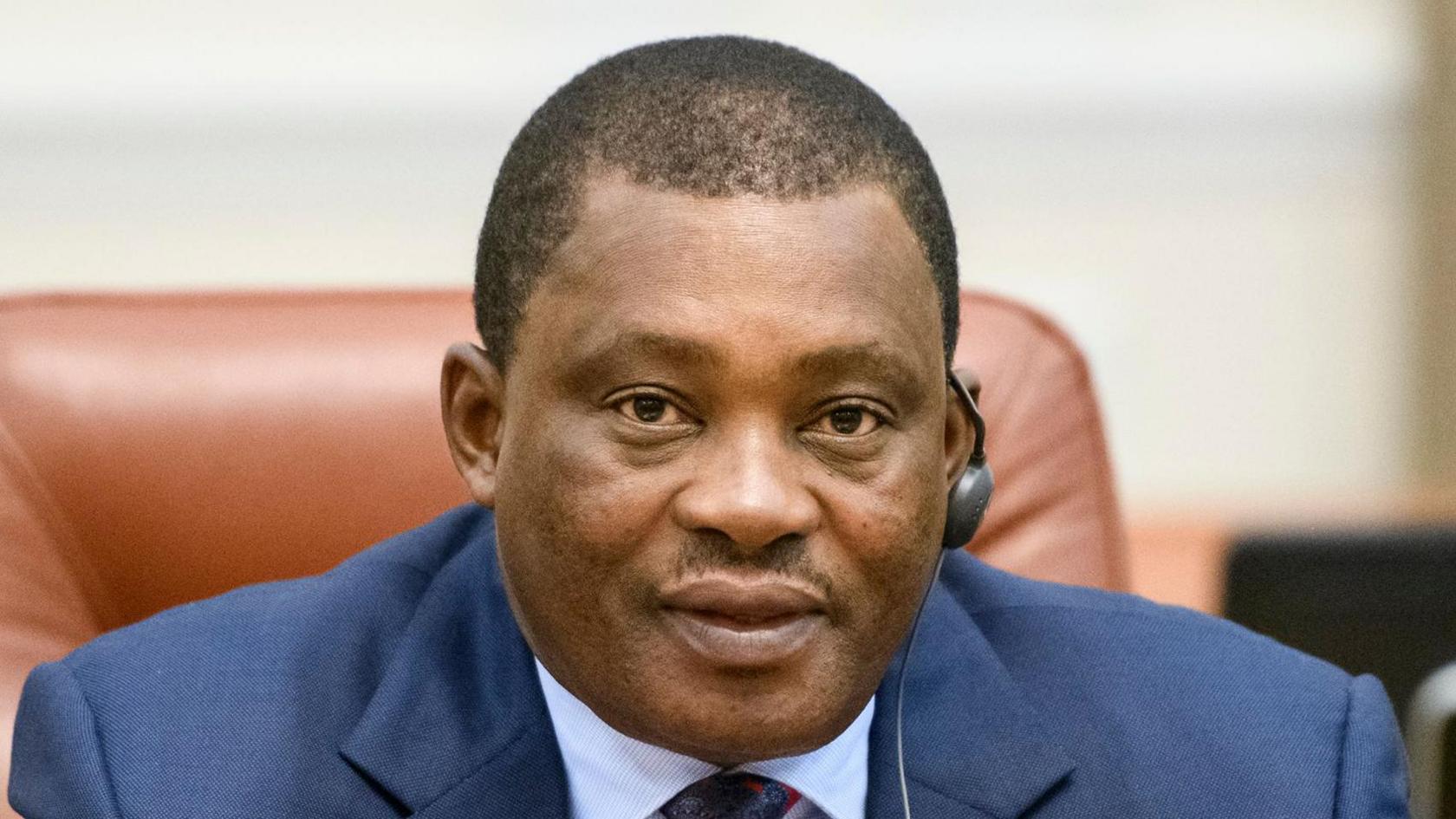 Public Service Cabinet Secretary Justin Muturi speaks in a microphone while seating on a leather chair. He is wearing a navy blue suit jacket and a light blue shirt and a multicoloured tie.