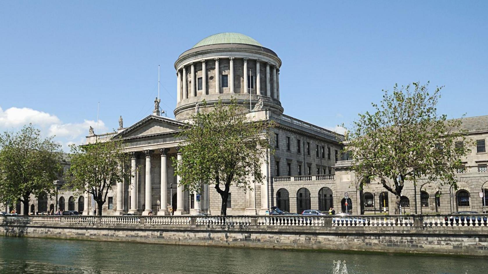 A wide shot of Dublin's High Court, showing a large white stone building with a large dome top. In front of the building is the River Liffey with four trees set up along the river's edge. The sky is blue and there are a few small clouds on the left hand side of the image.