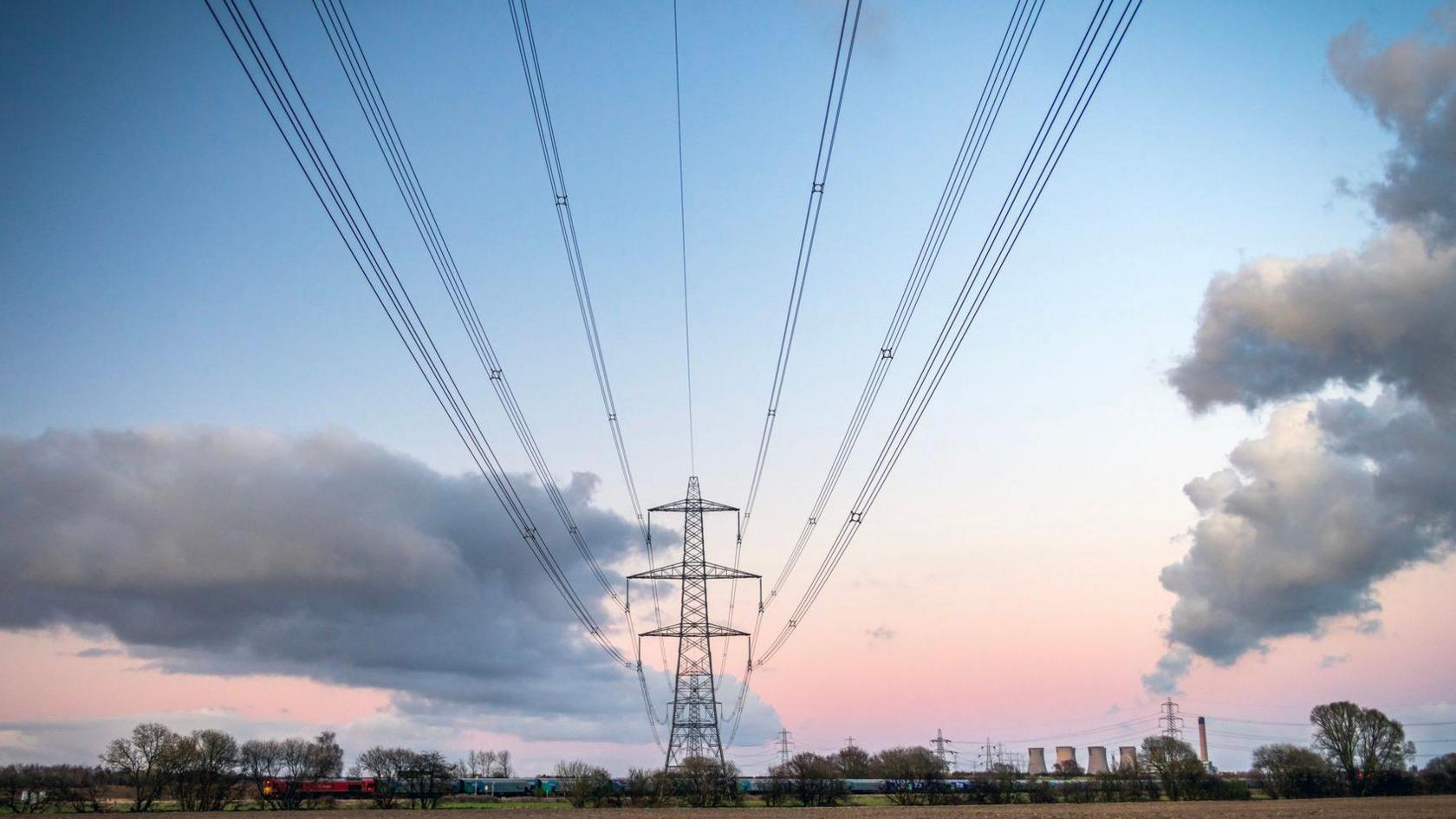 Electricity power lines going across a field with clouds and a ink and blue sky in the background.