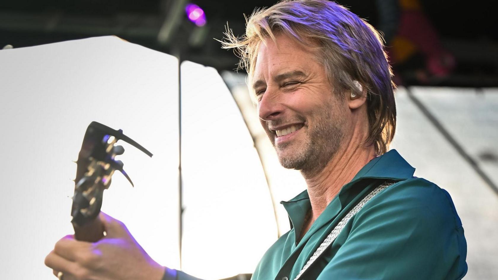 Singer Chesney Hawkes, with blonde hair and a green shirt, smiles and holds the neck of his guitar on stage