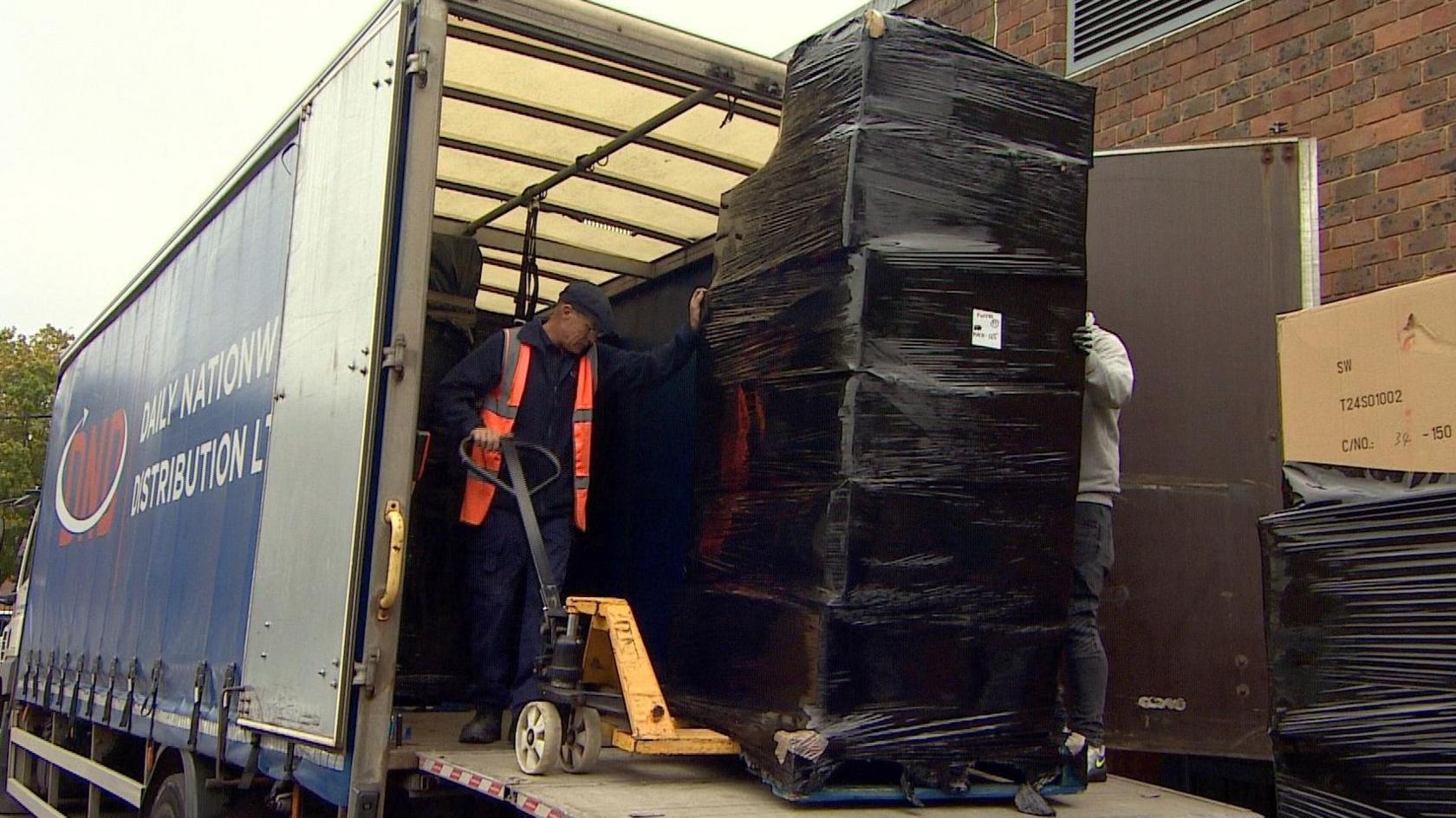 A pallet stacked with boxes covered in a black plastic wrap being unloaded from the back of a lorry. A man wearing a flat cap and high visibility jacket can be seen manoeuvring a hand-cranked forklift trolley under the pallet. Another man can just be seen behind the boxes wrapped in plastic. The cargo is stacked higher than both men and almost to the roof of the lorry. The side of the lorry can be seen on the left of the image, while in the foreground on the right is another stack of boxes wrapped in black plastic. 