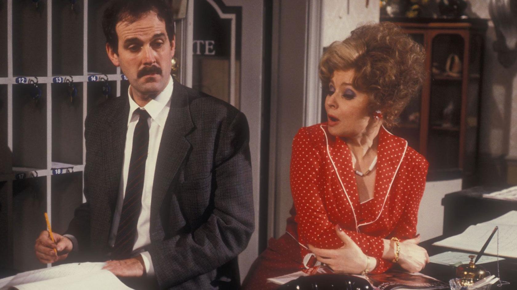 Prunella Scales in a red dress leaning on the reception desk as Sybil in Fawlty Towers and John Cleese as Basil