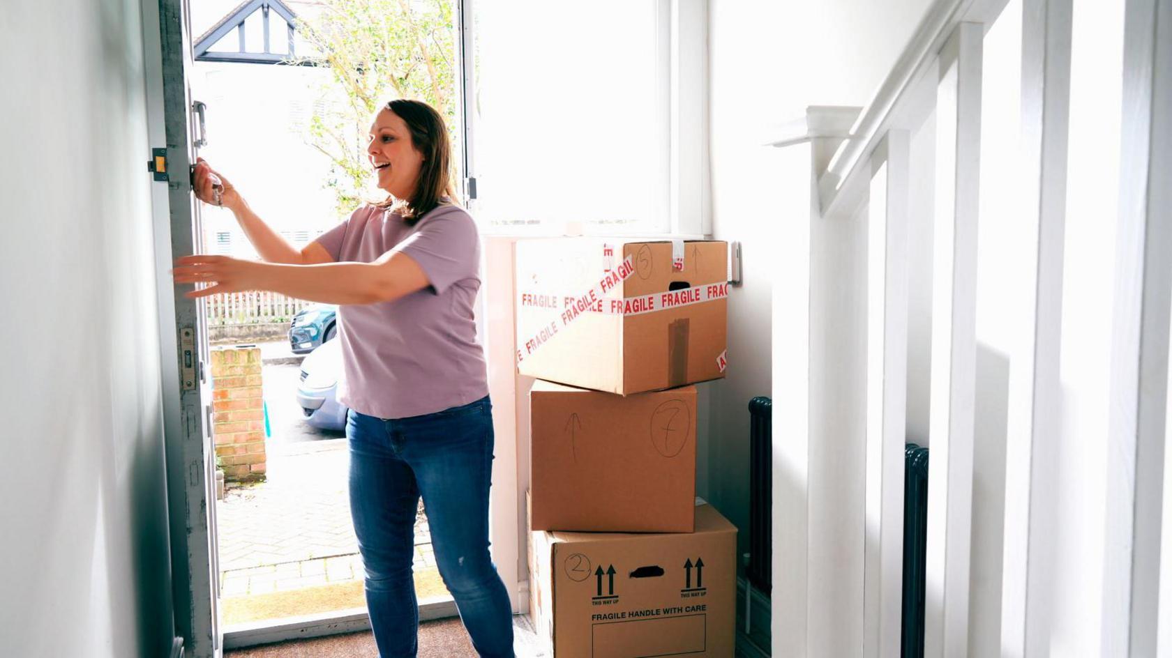 A woman moving into a new home. She is standing inside the house and is removing her keys from the front door. Three cardboard boxes are piled up behind her. The one on the top is closed with packing tape that reads, fragile.