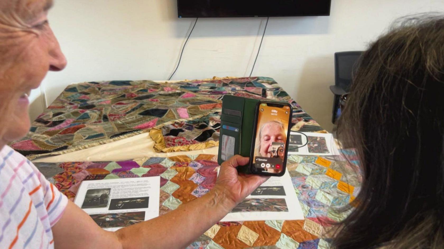 Two women on a video call with a third woman. In front of the group is a table with a patchwork quilt laid on top of it, as well as various bits of paper with words and pictures on them.