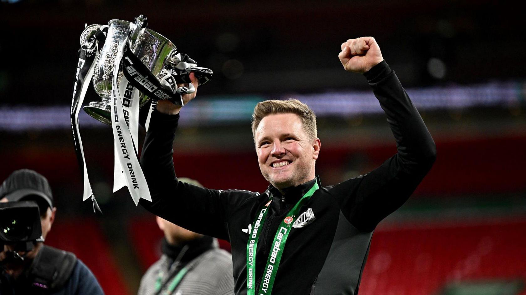 Eddie Howe holding the Carabao Cup aloft at Wembley. He has short fair hair and stubble and is wearing a black Newcastle United top. He is holding the silver trophy in his right hand. It is decorated with black-and-white ribbons.