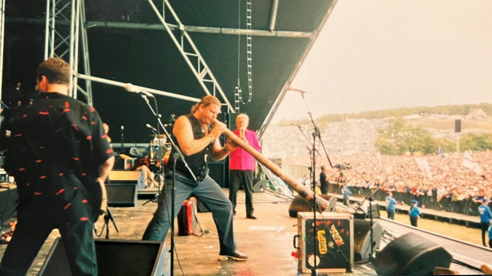 View is from the side of a stage with a crowd of thousands in front of it. A man in blue jeans and a dark coloured waistcoat is playing a didgeridoo. Rolf Harris, a man with greying hair, black trousers and a bright pink shirt, is standing behind him