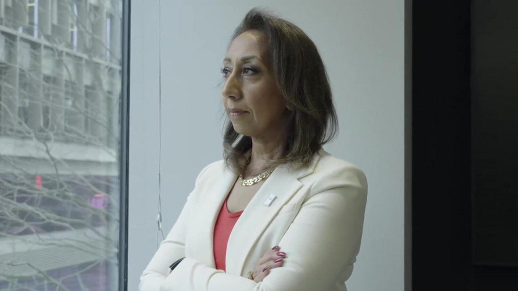 Moni Mannings, a woman with short brown hair wearing a cream jacket and coral top, looking out of the window of an office building with her arms folded