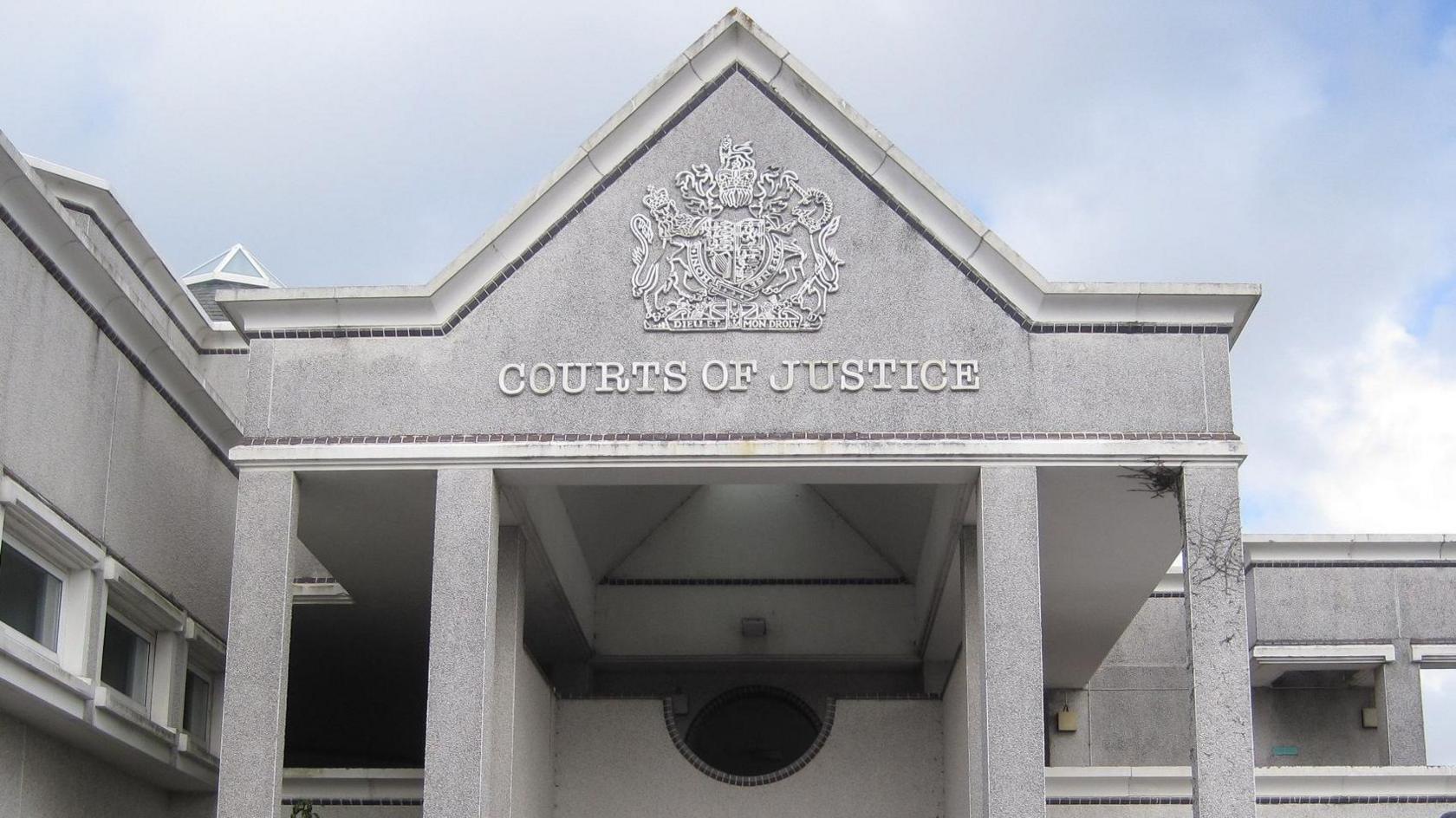 Truro Crown Court is pictured. It is made of pale grey granite and has four pillars and a steeple roof. There is an ornate heraldic shield on the front entrance and the words 'Courts of Justice'.