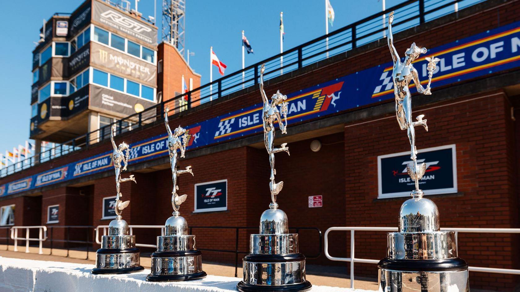 Large silver Classic TT trophies line up in front of the grandstand, which has TT  branding running along it.